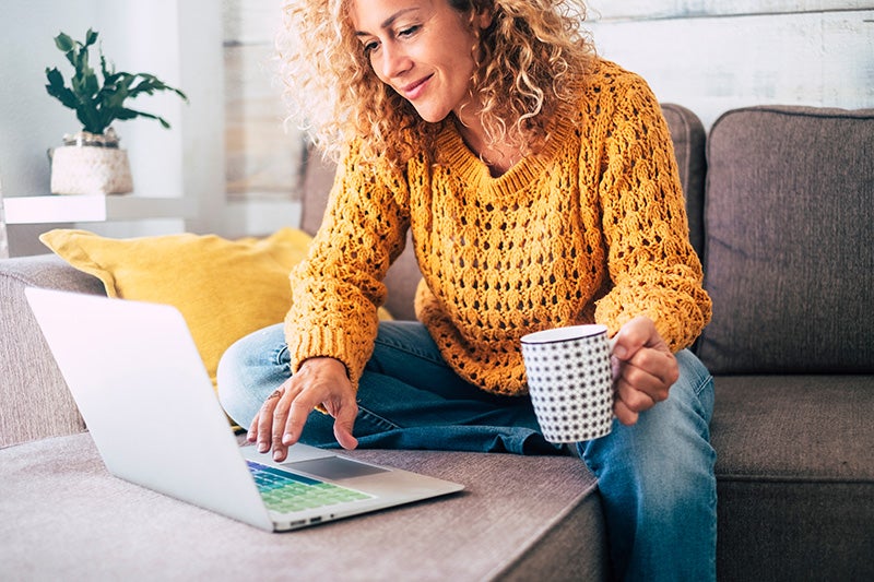 A woman, siting on a couch with a mug in one hand, while using a laptop to shop for a car